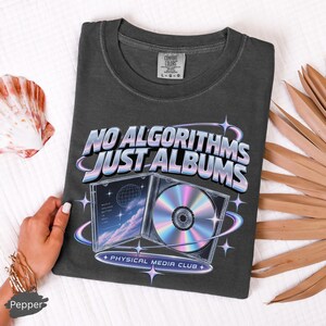 May include: A charcoal gray t-shirt featuring a graphic print with the text "NO ALGORITHMS JUST ALBUMS" and "PHYSICAL MEDIA CLUB". The design includes an open CD jewel case with a holographic disc and a sky illustration with a wireframe globe.