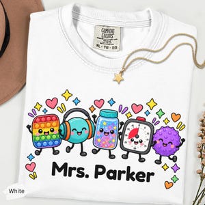 May include: A white t-shirt featuring a colorful graphic of five cartoon characters including a rainbow pop-it toy, headphones, a glitter jar, a kitchen timer, and a purple spiky ball, with the text Mrs. Parker printed in black below them.