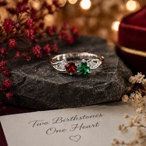 May include: A silver ring featuring a red and green gemstone design. The ring is displayed on a dark grey stone, with a card that reads "Two Birthstones One Heart". The background includes red flowers and a maroon jewellery box.