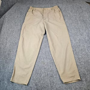 May include: A pair of khaki-colored trousers featuring an elasticized waistband, a front button closure, and a zippered fly. These pants have a relaxed fit with straight legs and side pockets, constructed from a solid-colored cotton-like fabric.