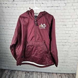 May include: A maroon quarter-zip pullover jacket with a hood and a front kangaroo pocket. The chest features the white Greek letters Alpha Phi with the text Alpha Phi printed underneath. The jacket has long sleeves and a drawstring at the hem.
