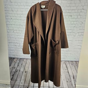 May include: A long, solid brown open-front coat featuring a large hood, wide lapels, and two prominent patch pockets on the front. The garment has long sleeves and a label at the neckline that reads 'Dales'.