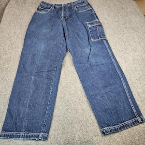 May include: Dark blue denim cargo pants featuring a relaxed fit, a button and zipper fly closure, belt loops, two front pockets, and a single rectangular cargo pocket on the left thigh with a small red and black brand label tag attached.