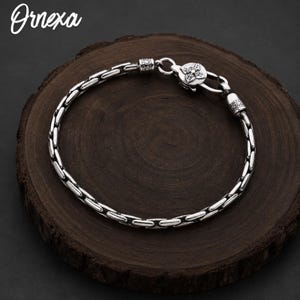 May include: A silver-toned chain bracelet featuring a series of elongated, rounded links and a circular clasp adorned with a four-petaled floral motif. The text Ornexa is visible in the upper left corner of the image.