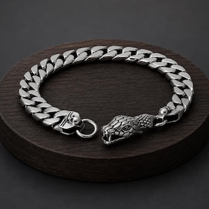 Könnte beinhalten: Ein Panzerkettenarmband aus silberfarbenem Metall mit einem detaillierten Schlangenkopf-Verschluss an einem Ende und einem kleinen Totenkopf-Emblem, das in die Gliederkonstruktion am anderen Ende integriert ist, sowie einem runden Ring zum Verschließen.