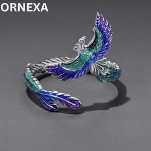 Peut inclure: Une bague ouverte argentée représentant un phénix avec des détails en émail bleu et vert vifs sur les ailes et les plumes de la queue. L'oiseau est représenté en plein vol avec les ailes déployées, et l'anneau s'enroule pour former la queue.