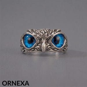 May include: A silver-toned metal ring shaped like an owl head, featuring detailed textured feather patterns and two prominent, vibrant blue glass eyes. The word ORNEXA is printed in black in the bottom left corner of the image frame.