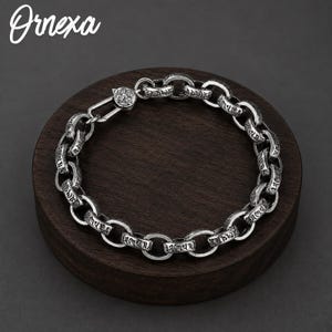 Könnte beinhalten: Ein silberfarbenes Kettenarmband mit dicken, abgerundeten Gliedern und eingravierter schwarzer Schrift. Das Armband verfügt über einen sicheren Karabinerverschluss. Das Wort Ornexa ist in der oberen linken Ecke des Bildes weiß gedruckt.