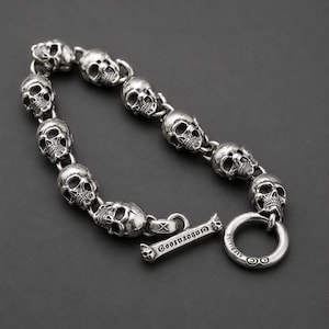 Op de afbeelding: Een zilverkleurige armband bestaande uit acht met elkaar verbonden schedelvormige schakels. De armband heeft een knevelsluiting met een staafje waarop de tekst "GOOTATOO" is gegraveerd en een ring met het woord "STERLING" en twee symbolen.