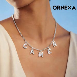Peut inclure: Collier argenté épelant "CAMEN" avec des cristaux clairs et étincelants. Les lettres sont suspendues à une chaîne délicate. Le mot "ORNEXA" est imprimé en noir en haut à droite.