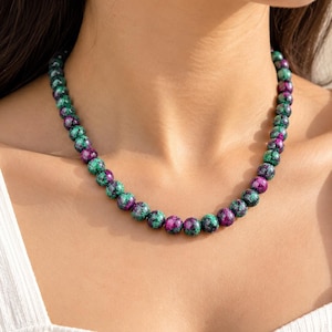 May include: A beaded necklace featuring round, polished stones in alternating shades of deep magenta and forest green. The spherical beads are strung together in a continuous strand, creating a vibrant, multi-coloured pattern around the neck.