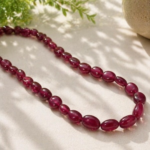 Puede incluir: Un collar con cuentas ovaladas de color rojo intenso. Las cuentas están ensartadas, creando un bucle continuo. El collar se muestra sobre una superficie clara, con un toque de vegetación en el fondo. Las cuentas tienen un aspecto pulido y brillante.