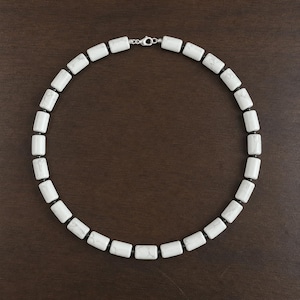 Puede incluir: Collar de cuentas blancas y negras con cierre plateado. El collar presenta cuentas rectangulares blancas con detalles negros, separadas por pequeñas cuentas negras. El collar se muestra sobre una superficie marrón oscuro.