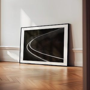 May include: Framed black and white photograph of a winding road with white lines. The artwork is leaning against a white wall on a wooden floor, creating a minimalist aesthetic.