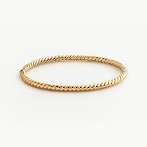 Może przedstawiać: Złota bransoletka typu bangle w stylu skręconej liny. Bransoletka ma ciągły, okrągły wzór z teksturowaną powierzchnią. Biżuteria jest prezentowana na białym tle.