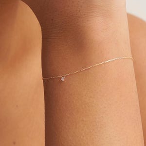 Op de afbeelding: Een delicate gouden armband met een kleine, sprankelende diamant hanger. De armband is dun en minimalistisch, ontworpen om op de arm te worden gedragen. De diamant is een kleine, heldere steen, die een vleugje elegantie aan de sieraden toevoegt.