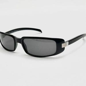 May include: Black rectangular sunglasses with dark gray lenses. The sunglasses have a sleek, modern design with a silver accent on the temples. The frame is made of a glossy black material.