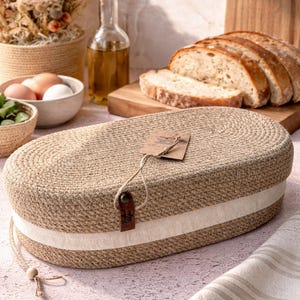 Puede incluir: Cesta ovalada tejida en beige natural, con una franja blanca. Una etiqueta está sujeta con una correa de cuero. Ideal para guardar pan o para uso en la cocina. Mide unos 30 cm de largo.