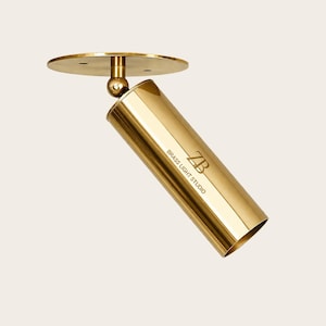 May include: A gold-colored, cylindrical spotlight fixture with a circular base and adjustable ball joint. The fixture has the text "BRASS LIGHT STUDIO" and a "ZB" logo. The spotlight is designed for ceiling mounting.