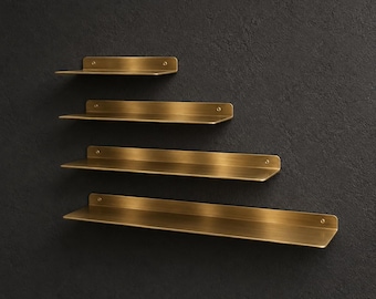 May include: A set of four wall-mounted shelves made of brushed brass metal. Each shelf features a flat horizontal surface and a vertical back panel with small circular screw holes for mounting, arranged in a staggered, ascending length pattern.