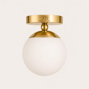 May include: A modern ceiling light fixture with a gold finish and a white globe shade. The fixture has a round base with the text "ZB BRASS LIGHT STUDIO" and a spherical glass shade. The overall design is minimalist and elegant.