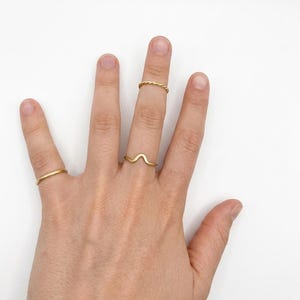 Puede incluir: Una mano muestra cuatro anillos dorados sobre un fondo blanco. Los anillos incluyen una banda fina, una banda trenzada y un anillo en forma de onda. Los anillos son simples y elegantes, adecuados para el uso diario.