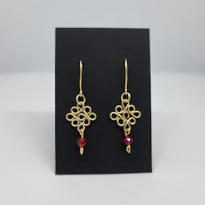 Puede incluir: Un par de pendientes dorados con un diseño de nudo decorativo. Cada pendiente tiene una pequeña cuenta roja colgante. Los pendientes se exhiben en una tarjeta negra sobre un fondo blanco.