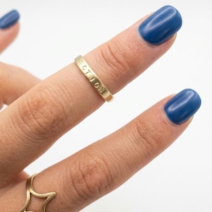 Puede incluir: Un anillo dorado con la palabra "ACTION" grabada se lleva en un dedo con esmalte de uñas azul. También es visible otro anillo dorado con un diseño en forma de estrella. El fondo es blanco.
