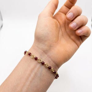 Puede incluir: Un brazalete dorado con cuentas rojas y detalles en espiral. El brazalete se muestra en una muñeca sobre un fondo blanco. Las cuentas son pequeñas y redondas, y el oro tiene un acabado brillante.