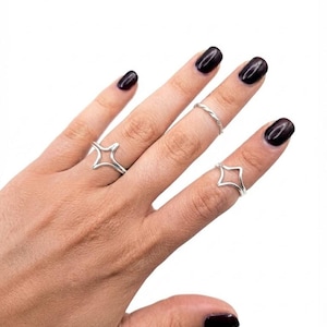 Puede incluir: Tres anillos de tono plateado usados en los dedos. Dos anillos presentan un diseño abierto en forma de estrella de cuatro puntas o diamante, mientras que el tercer anillo es una banda simple, delgada y trenzada. Joyería metálica pulida.