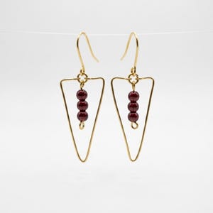 Puede incluir: Un par de pendientes dorados con un diseño triangular. Cada pendiente presenta tres cuentas de color rojo oscuro suspendidas dentro del triángulo. Los pendientes tienen un diseño simple y elegante, adecuado para el uso diario.