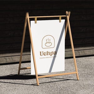 Może przedstawiać: Drewniany szyld w kształcie litery A z białym prostokątnym panelem. Na szyldzie widnieje brązowe logo i nazwa "Uchpa" z ilustracją chleba. Szyld stoi na zewnątrz na szarej powierzchni.