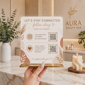 May include: A clear acrylic sign with a gold metal base featuring the text LET'S STAY CONNECTED, FOLLOW ALONG, SCAN TO CONNECT, FOLLOW OUR JOURNEY @aurabeautybar, LOVE YOUR EXPERIENCE? LEAVE US A REVIEW, and we appreciate you! with two QR codes.