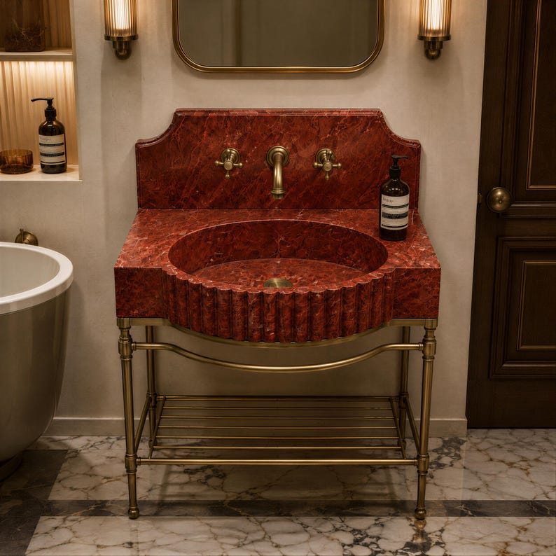 a bathroom with a red marble sink and a gold-framed mirror above it. The sink has a unique, textured design and is placed on a metal stand. The bathroom also includes a bathtub and a door, creating a well-appointed and elegant space.