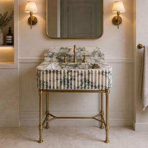 Puede incluir: Un lavabo de baño de mármol blanco y gris con un diseño exterior acanalado, montado sobre un soporte de metal de latón. El lavabo cuenta con un protector contra salpicaduras de mármol a juego, un grifo central y dos manijas de latón.
