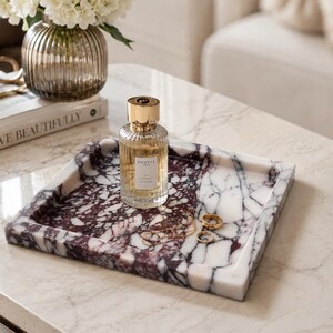 May include: A square marble tray featuring a deep burgundy and white veined pattern. The tray holds a clear glass perfume bottle with a gold cap labeled Goutal Paris, along with a thin gold chain necklace and two small gold rings resting inside.