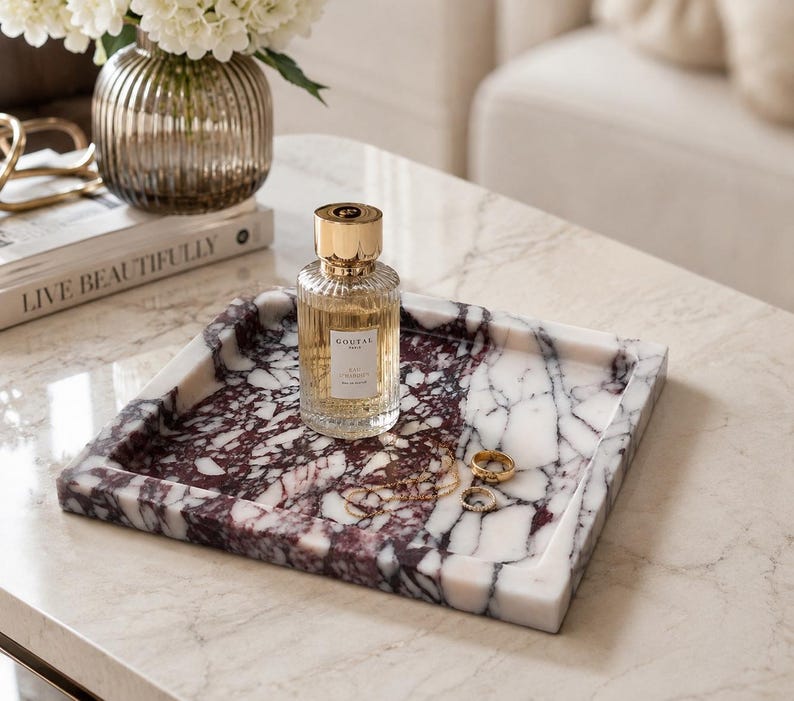 May include: A square marble tray featuring a deep burgundy and white veined pattern. The tray holds a clear glass perfume bottle with a gold cap labeled Goutal Paris, along with a thin gold chain necklace and two small gold rings resting inside.