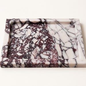 May include: A square tray crafted from polished stone featuring a distinct brecciated pattern. The surface displays a high-contrast mix of deep burgundy and stark white angular fragments, creating a complex, fractured appearance with raised edges.