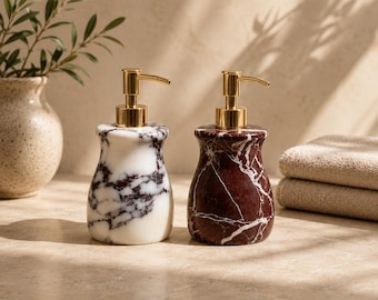 May include: Two marble soap dispensers with gold-colored pumps. One is white with dark gray veining, the other is dark red with white veining. The dispensers are on a light-colored surface, with a vase and plant in the background.