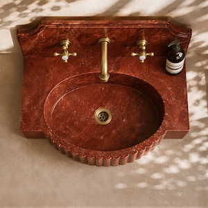 a red marble sink with a gold faucet, placed on a beige tiled floor. There is a bottle of soap next to the sink.