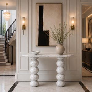 May include: A white marble console table with an oval top and two legs composed of stacked spherical marble spheres. A round beige ceramic vase containing green branches and a small matching marble bowl sit on the table surface.