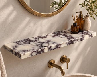 May include: A rectangular wall-mounted shelf featuring a white and dark purple marble pattern. The shelf is positioned above a brass faucet and a stone basin, with a glass bottle, a reed diffuser, and a small vase with greenery on top.