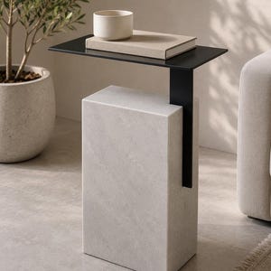 Pode incluir: Uma mesa lateral com uma base retangular de mármore branco e um tampo fino de metal preto que se estende horizontalmente. Um livro bege e uma pequena tigela redonda de cerâmica branca estão colocados sobre a superfície de metal.