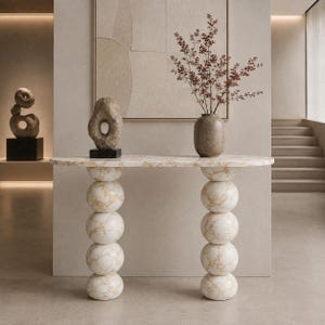 Op de afbeelding: Een consoletafel met een glad, ovaalvormig blad en twee verticale steunen, elk bestaande uit vier gestapelde witte marmeren bollen met gouden aders. Het tafeloppervlak komt overeen met het marmerpatroon van de bolvormige poten.
