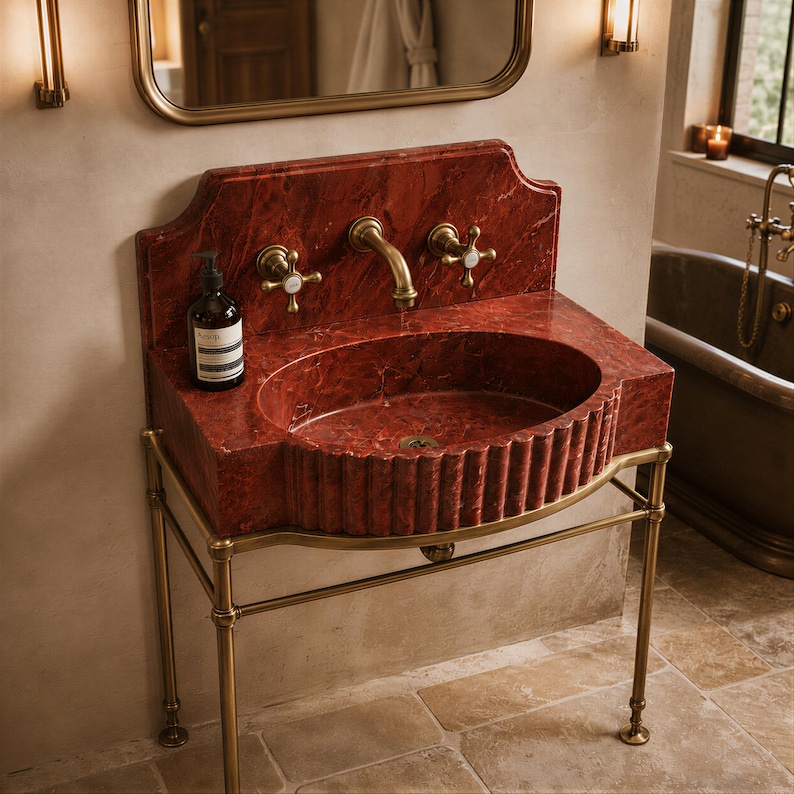 a bathroom with a red marble sink and a gold-framed mirror above it. The sink has a curved design and is placed on a metal stand.