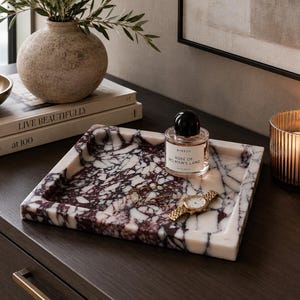 Puede incluir: Una bandeja cuadrada de mármol con base blanca y vetas de color púrpura oscuro y negro. Sobre la bandeja hay un frasco de perfume de vidrio transparente con tapón negro etiquetado Byredo Rose of No Man's Land y un reloj de pulsera de metal dorado.