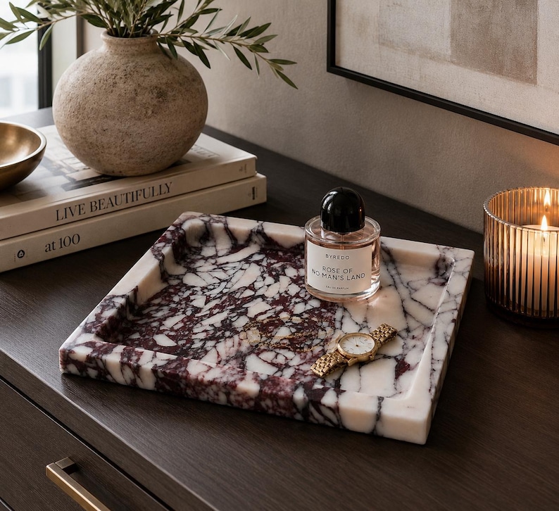 May include: A square marble tray featuring a white base with deep purple and black veining. Resting on the tray is a clear glass perfume bottle with a black cap labeled Byredo Rose of No Man's Land and a gold-toned metal wristwatch with a link band.