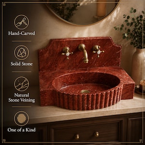 a bathroom sink made of red marble, with a round basin and two gold faucets. The sink is placed on a wooden cabinet, and there is a round mirror above it. The sink is hand-carved and made of solid stone, giving it a natural stone veining appearance. The sink is described as one of a kind, and it is described as having a hand-carved design and a natural stone veining pattern.