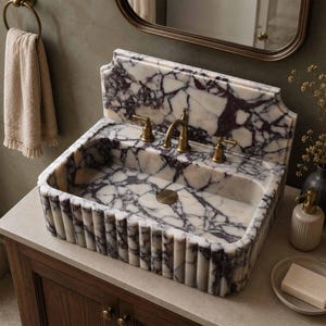 Puede incluir: Un lavabo rectangular de mármol blanco con vetas moradas, que presenta un diseño exterior acanalado y un salpicadero alto. El lavabo está equipado con un grifo central de latón y dos manijas de latón a juego sobre una encimera de piedra.