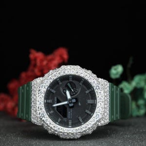 May include: A dark green analog-digital wristwatch featuring a round bezel encrusted with numerous clear, sparkling stones. The watch face is black with silver-toned hands and markers, and it includes a matching dark green resin strap.